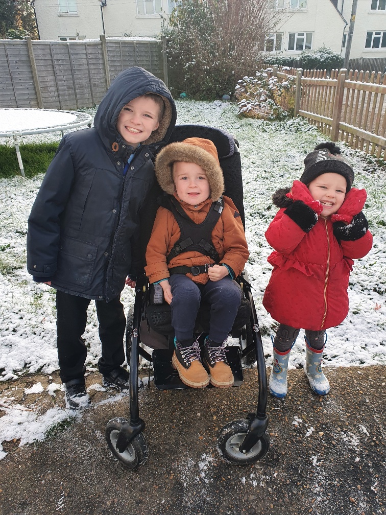 Three siblings wrapped up in coats in the snow. The middle child is in a wheelchair and has a brother and sister on either side.