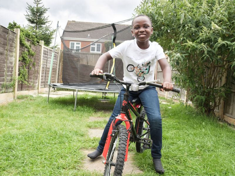 A boy riding a red bicycle in a garden with a trampoline behind him