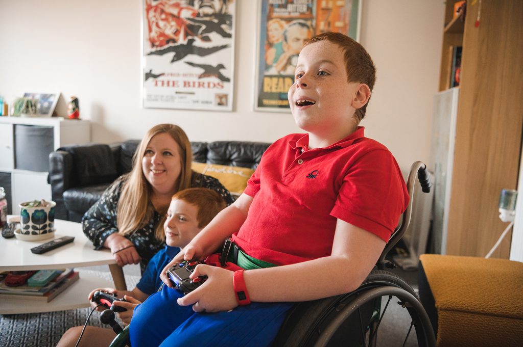 Boy in wheelchair plays with his brother on the PlayStation with mum watching on. 