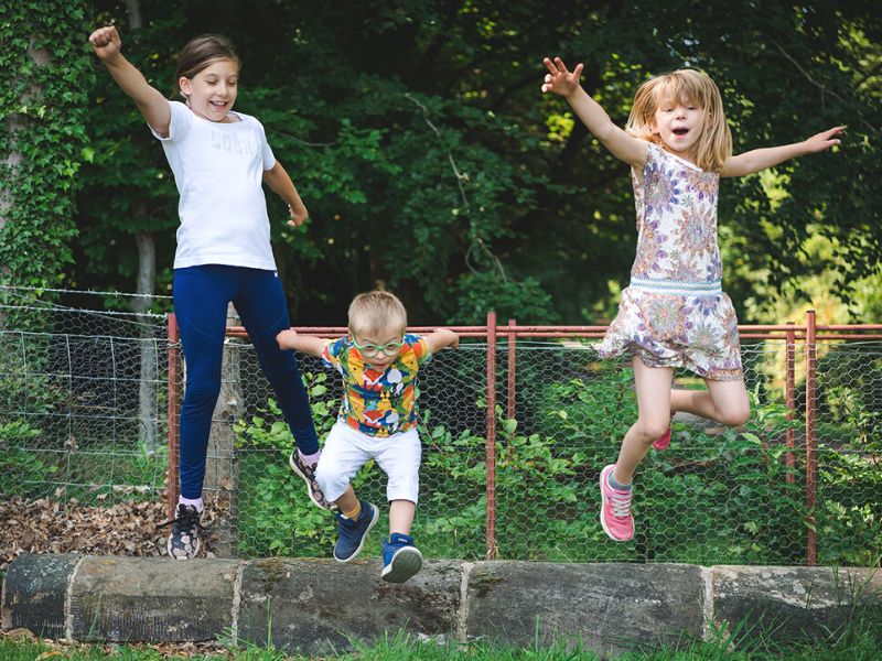 Three siblings, two girls and a boy in the middle, playing outside jumping into the air with their arms up
