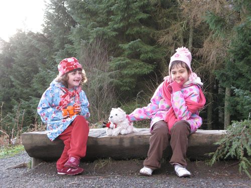 Two sisters sit on a bench in a forest wearing coats and hats