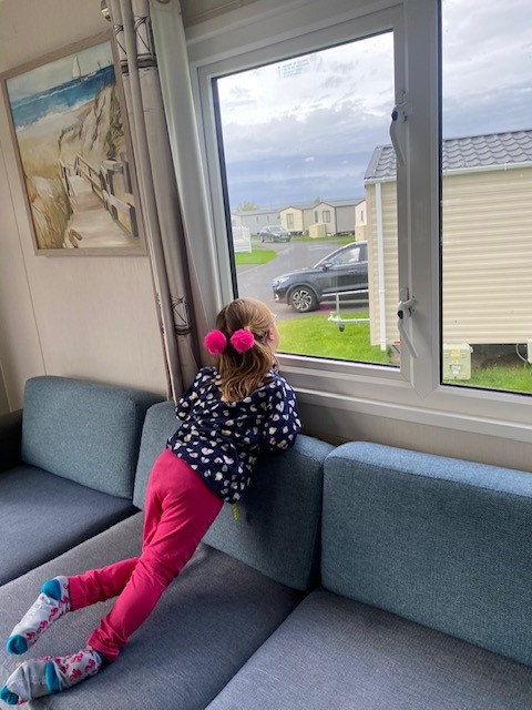 Little girl sits on sofa looking out of caravan window onto other caravans