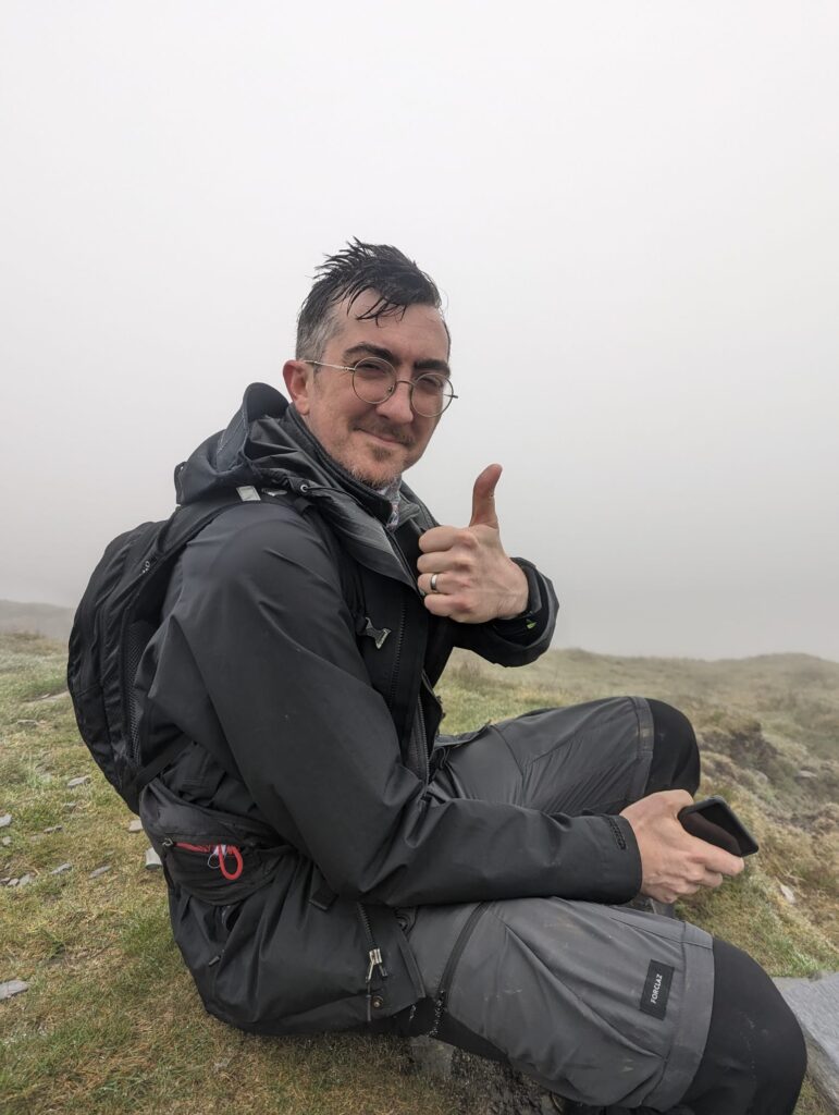 A man sat down at the top of a mountain, in the fog, giving a thumbs up to the camera 