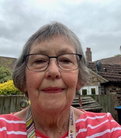 A woman with glasses and grey hair, wearing a pink striped shirt and a family fund lanyard is smiling at the camera