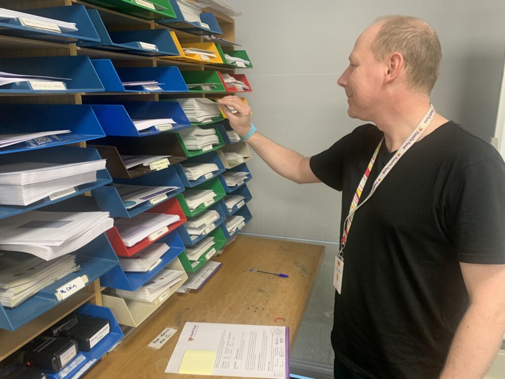 A man, wearing a black tee-shirt and a family fund lanyard, is filing letters in a large and colourful file cabinet
