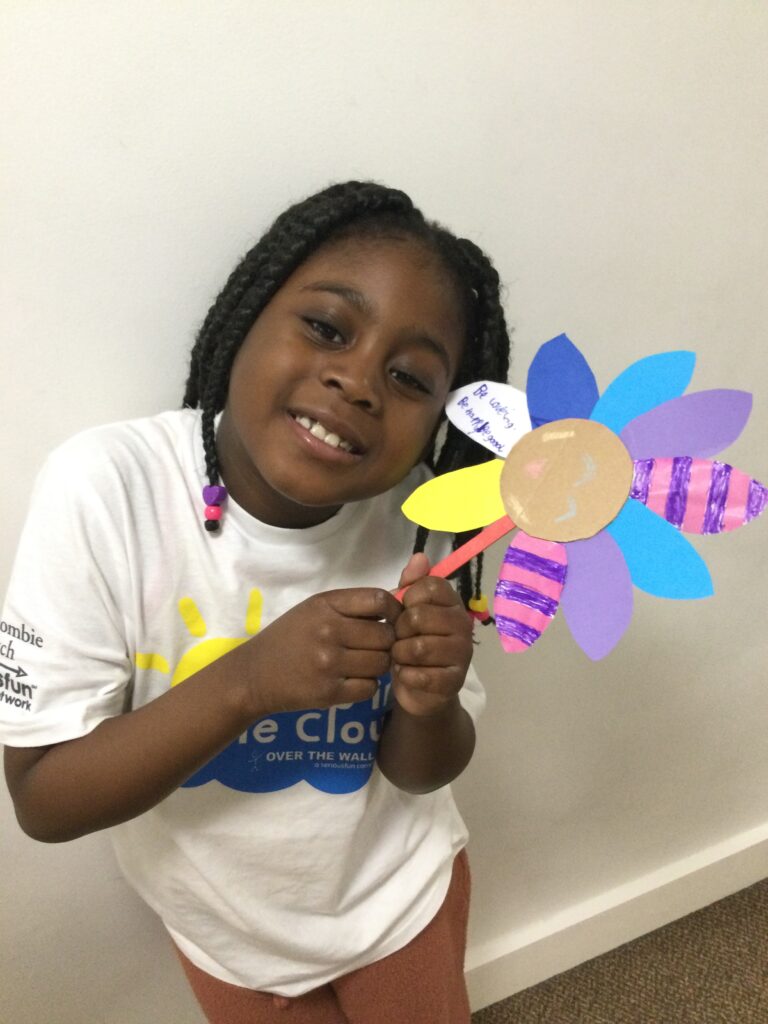 A young girl with a Camp in the Cloud tee-shirt is smiling at the camera, posing with a big paper flower she just crafted 