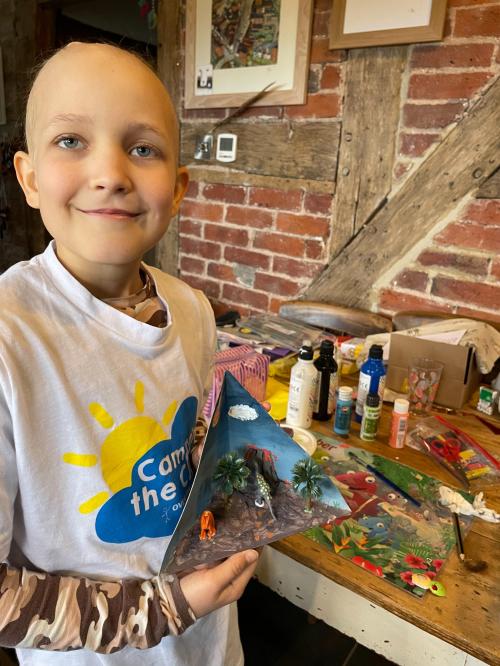 A child with a Camp in the Cloud tee-shirt is smiling at the camera, posing with some craft they just finished