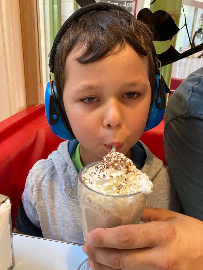 A boy with dark hair and pale eyes is wearing blue ear defenders and is enjoying a hot chocolate.