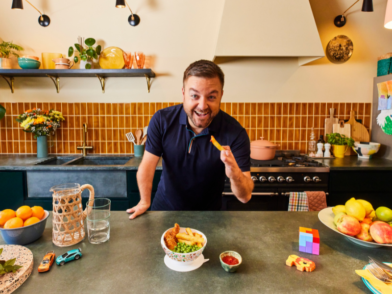 Comedian Alex Brooker stood in a kitchen holding the McCain and Family Fund scoop bowl in one hand and a spatula with chips on in the other