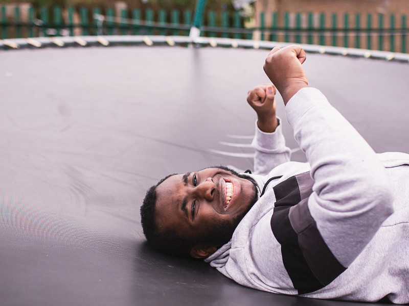 A teenager lies on a trampoline. He is smiling and his hands are raised in the air.