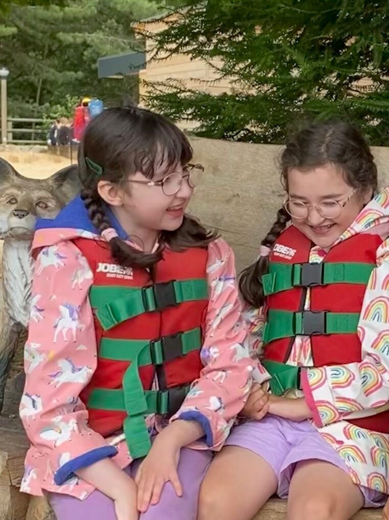 Sisters Ruby, aged nine, and Laura, aged ten, sit together on an outdoor bench. They are smiling and are wearing life jackets.