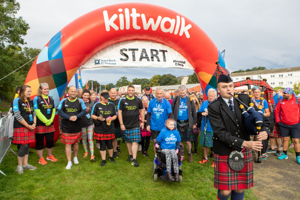A large red arch banner with 'Kiltwalk start' on it and in front are a large group of people wearing kilts being led by a man playing the bagpipes