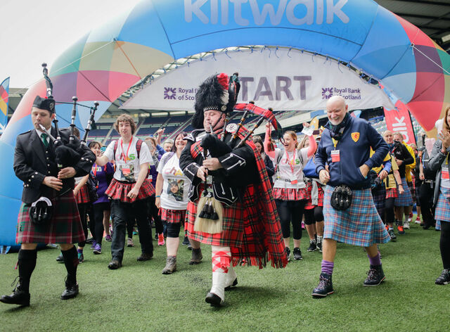A large arch banner with 'Kiltwalk' on it and in front are a group of people all wearing tartan kilts walking across the grass