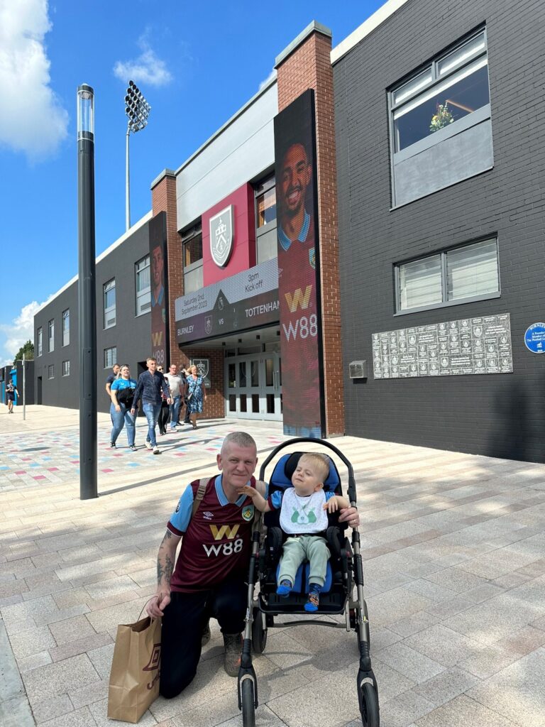 A young blond boy is sat in a stroller, outside a stadium. Next to him is a man wearing a football jersey, kneeling down next to the stroller, smiling at the camera