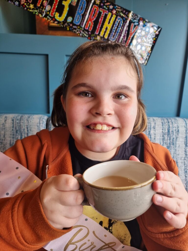 Ophelia, a 13-year-old girl, sits on a sofa, smiling at the camera as she drinks a cup of tea.