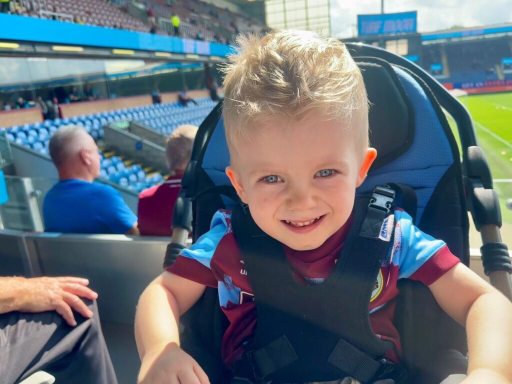 A young blond boy with blue eye is sat outside in a stroller, in the middle of a stadium