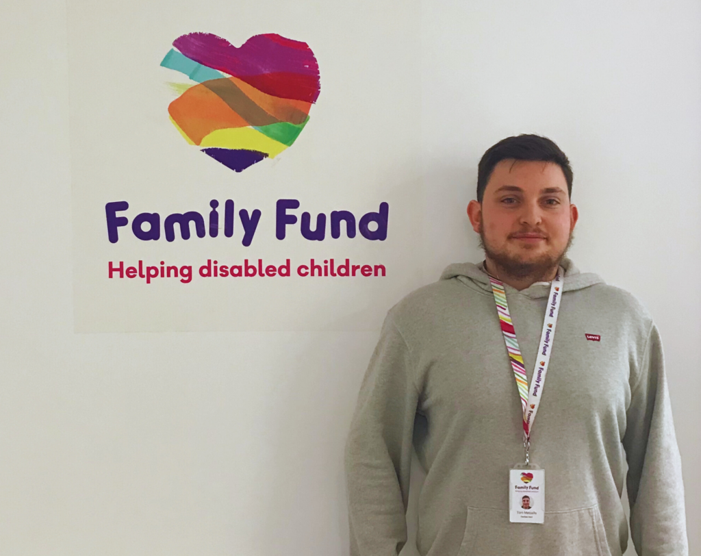 A young man with dark hair and wearing a grey hoodie and family fund lanyard poses, in front of a white wall with the family fund logo behind him