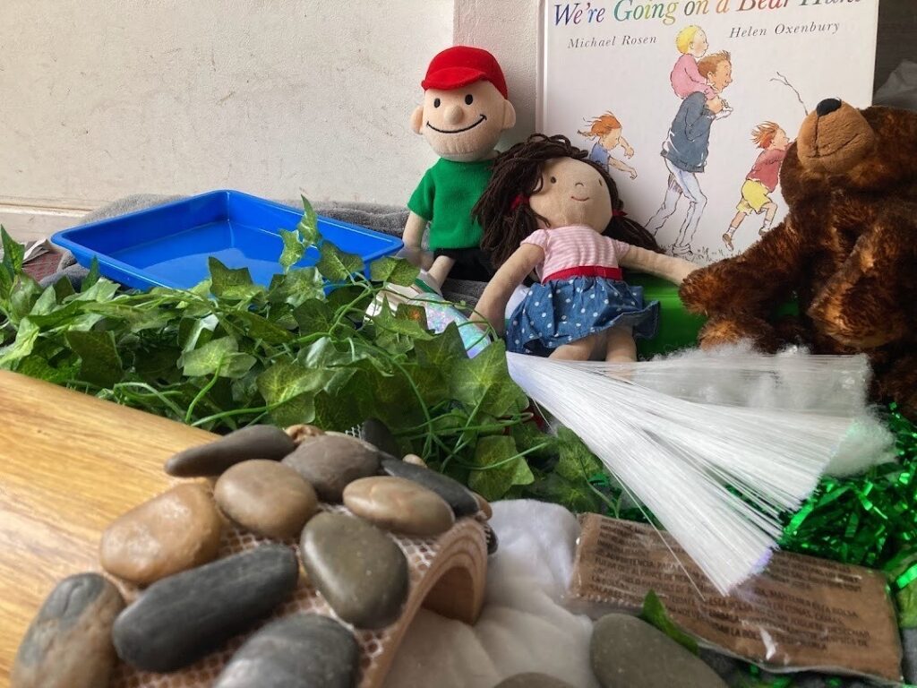 Example of a sensory story with items gathered next to the book 'We're going on a bear hunt' including pebbles, leaves, dolls, and a soft bear