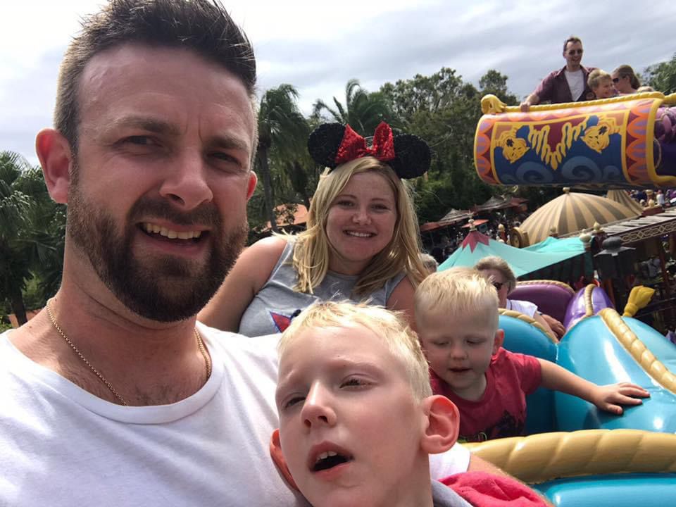 Kris, Jess, Archie and Mason are on a ride at Disney, Florida. Mason is next to Kris, with his head resting on his chest. Kris is smiling at the camera, and Mason is looking at the camera. Jess sits behind them smiling at the camera, wearing Minnie Mouse ears, witht Archie by her side.