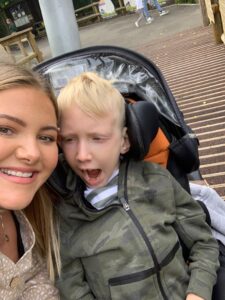Mason and Jess are outside. Mason is in a specialist pram, and Jess is taking the photo beside him with her face pressed against his.