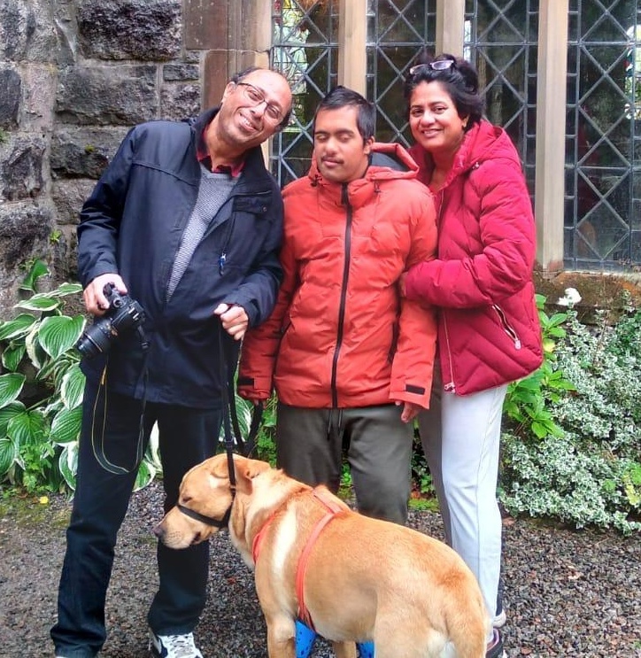 Aaryan is pictured with his mum and dad and their labrador. They are outdoors in front of an old building. Pronita and Subhayu are smiling, linking up with Aaryan.