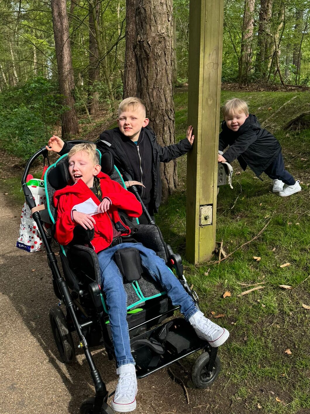 Mason and his brothers are surrounded by woodland. Archie is pushing Mason in his wheelchair, and Arthur is leaning against a nearby pole.