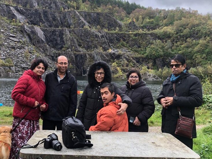 Aaryan, his grandparents, parents and brother are standing and sitting by a bench with a lake in the background. They are looking and smiling at the camera. They are pictured in the Highlands, Scotland.