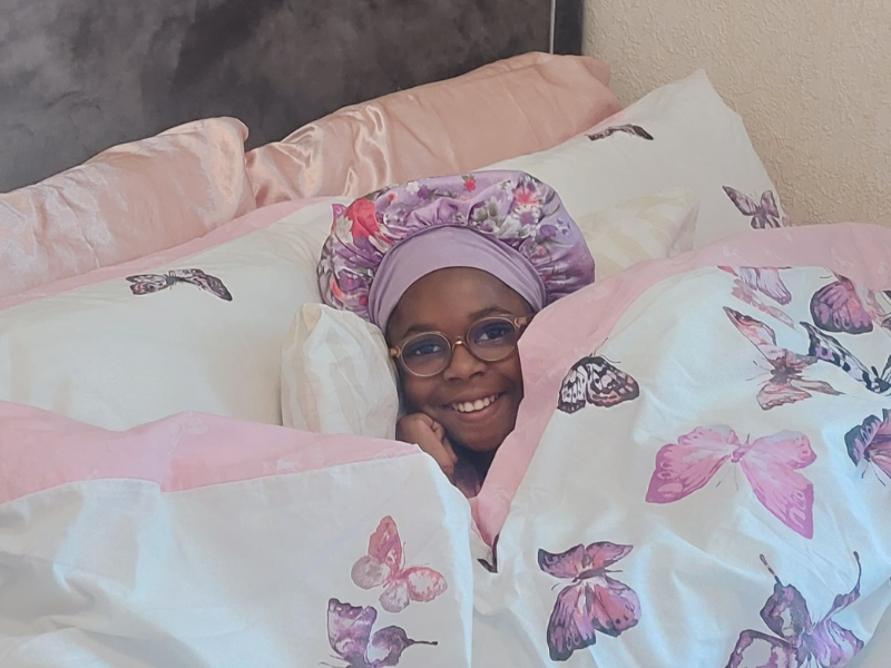 A little girl wearing her sleep cap lays in a cosy bed with white and pink bedding on
