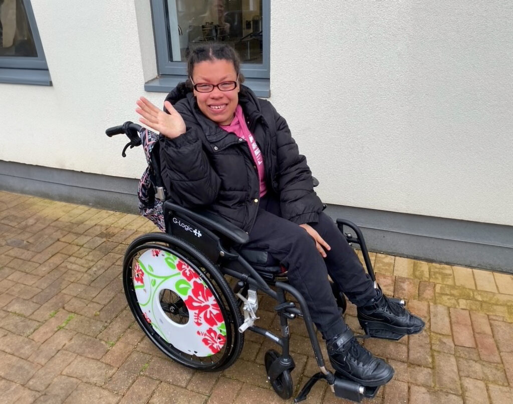 Nur, a young woman, wearing glasses and  outside in a wheelchair waving happily.