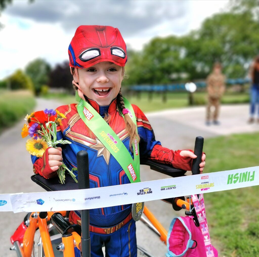 A young girl in a Captain Marvel outfit holding a bunch of flowers at a finishing line.
