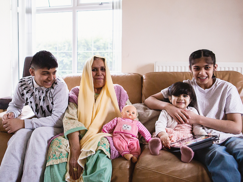 A woman is sat on the sofa next to her children: a teenage boy, a teenage girls and a young girl holding an iPad. They are smiling.
