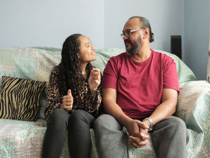A teenage girl is sat on the sofa next to her father. They are looking at each other and smiling.