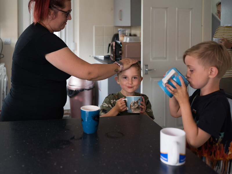 A woman is stroking her son's hair in the kitchen. He is holding a cup and his brother is having a drink next to him.