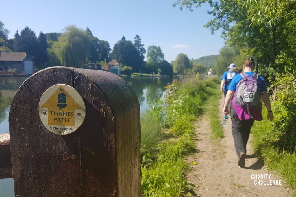 Walkers walking along a footpath next to a river. In the foreground is a post which is marked 'Thames Path' with an ahead arrow. 