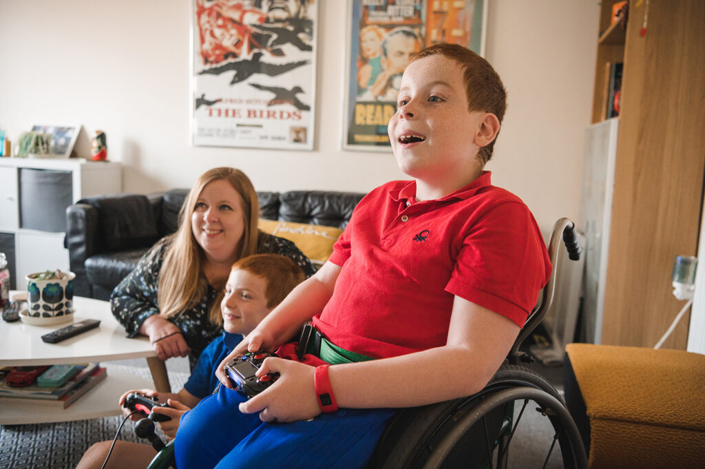 A young boy in a wheelchair plays on a games console with his younger brother, whilst mum watches on smiling.