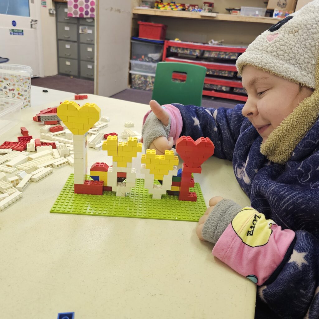 A young girl in a hat, gloves and cosy hoodie is looking at her lego creation, giving a thumbs up to the camera.