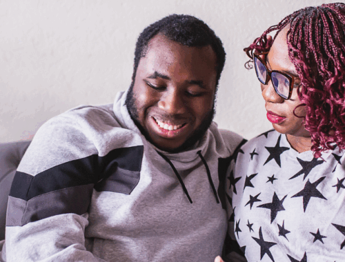 A young adult son smiling, sat on a sofa next to his mum wearing glasses and a starred t-shirt
