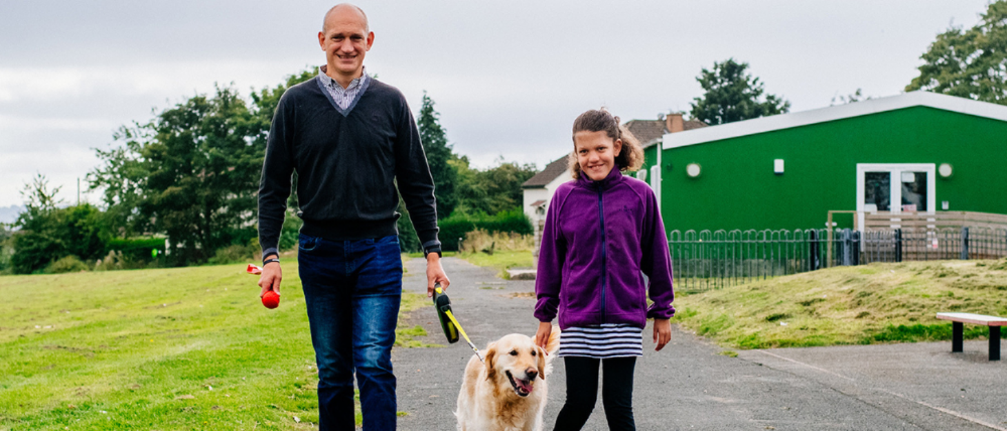 A photo of a dad walking with his daughter and dog in the park.