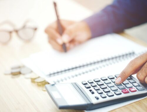 A close up of a desk area. A person is holding a pen in their right hand over a notepad, and using the calculator with their left hand.