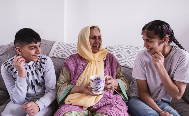 A photo of a woman sat on the sofa holding a mug. Next to her are a teenage boy and girl wearing hearing aids. They are looking at her and smiling.