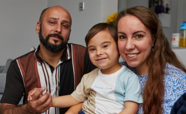 A photo of a man and woman holding their son. They are all smiling at the camera.