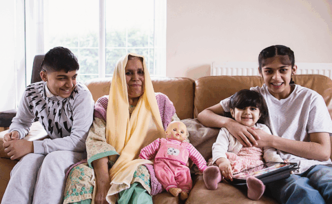 A woman is sat on the sofa next to her children: a teenage boy, a teenage girls and a young girl holding an iPad. They are smiling.