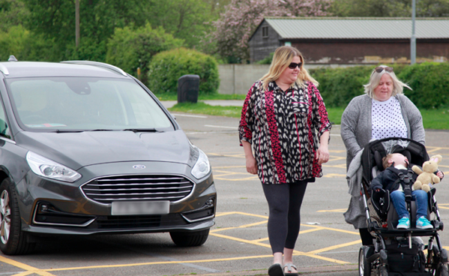 Two women are walking away from their car in the car park. The older woman is pushing a pram where a young boy is sat.