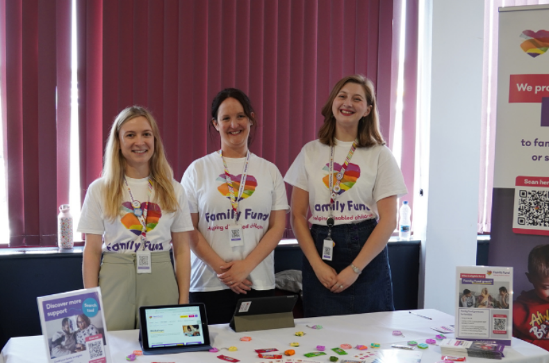 Three young women wearing Family Fund branded t-shirts next to the stall are smiling at the camera.