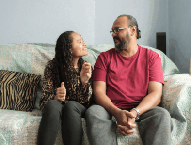 A teenage girl is sat on the sofa next to her father. They are looking at each other and smiling.