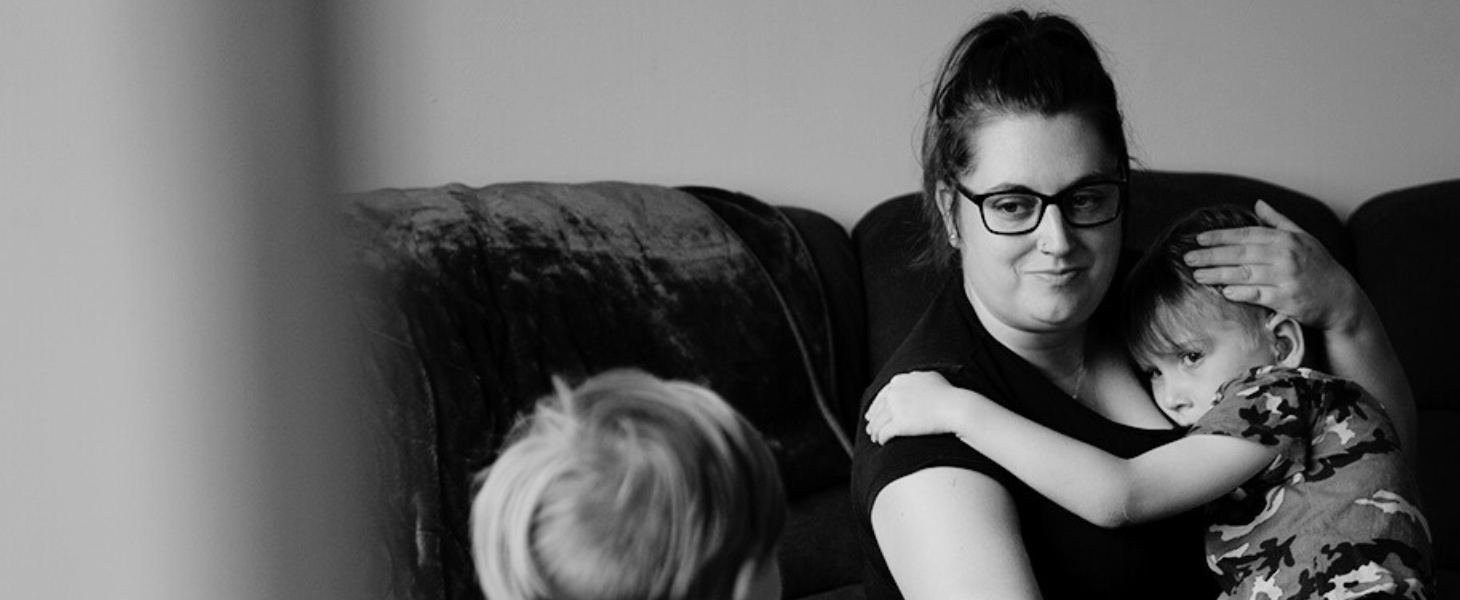 Black and white photo of a mum sat on a sofa cuddling her son looking toward her other son