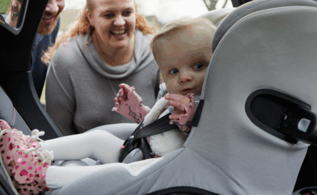 Olivia sits in her car seat in the back of their Family Fund Motability Programme lease car and her mum and dad smile at her through the open car door.