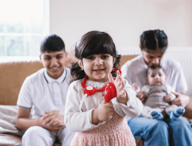 A child with a plastic stethoscope smiling. Two children are sat on a sofa in the background smiling, one of them with a baby on her lap.