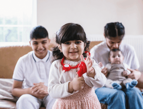 A child with a plastic stethoscope smiling. Two children are sat on a sofa in the background smiling, one of them with a baby on her lap.