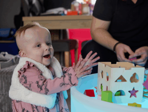 A young girl smiling and clapping her hands in front of a block toy.